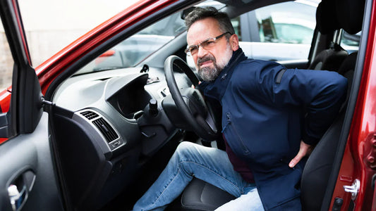Side view of a man with glasses and a blue jacket sitting in a car, looking distressed and holding his lumbar spine. The image highlights how car seats can contribute to back discomfort